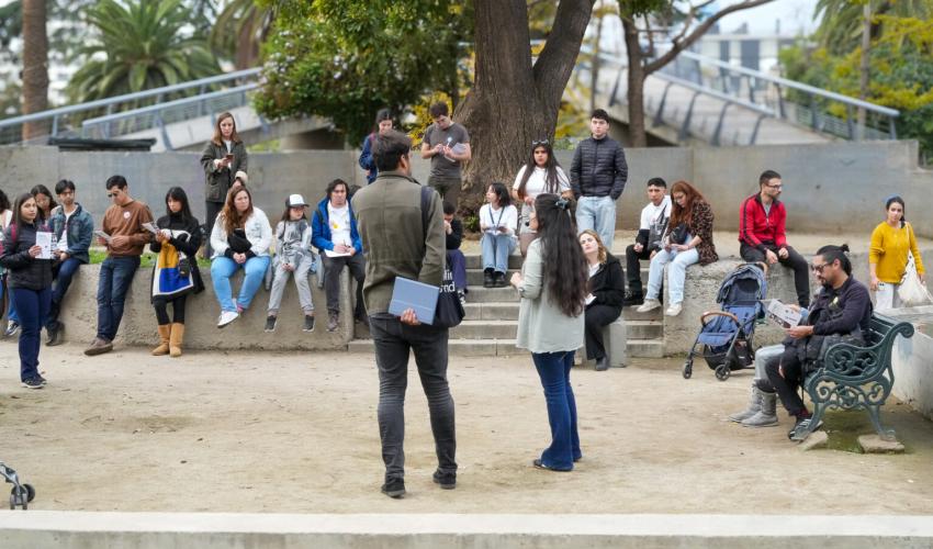 Imagen de un grupo de personas reunidas en el espacio público con dos mediadores que explican la historia y arquitectura del lugar.