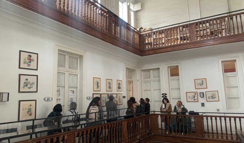 Vista interior de la Casa Maroto durante una visita guiada. La imagen muestra un amplio espacio de doble altura con paredes blancas decoradas con cuadros y grabados. Un grupo de visitantes recorre un pasillo en el segundo nivel, delimitado por una elegante balaustrada de madera tallada que rodea el tragaluz central. La luz natural entra por ventanas altas en el nivel superior, destacando los detalles arquitectónicos clásicos y la atmósfera cultural del Club de Jazz de Santiago.